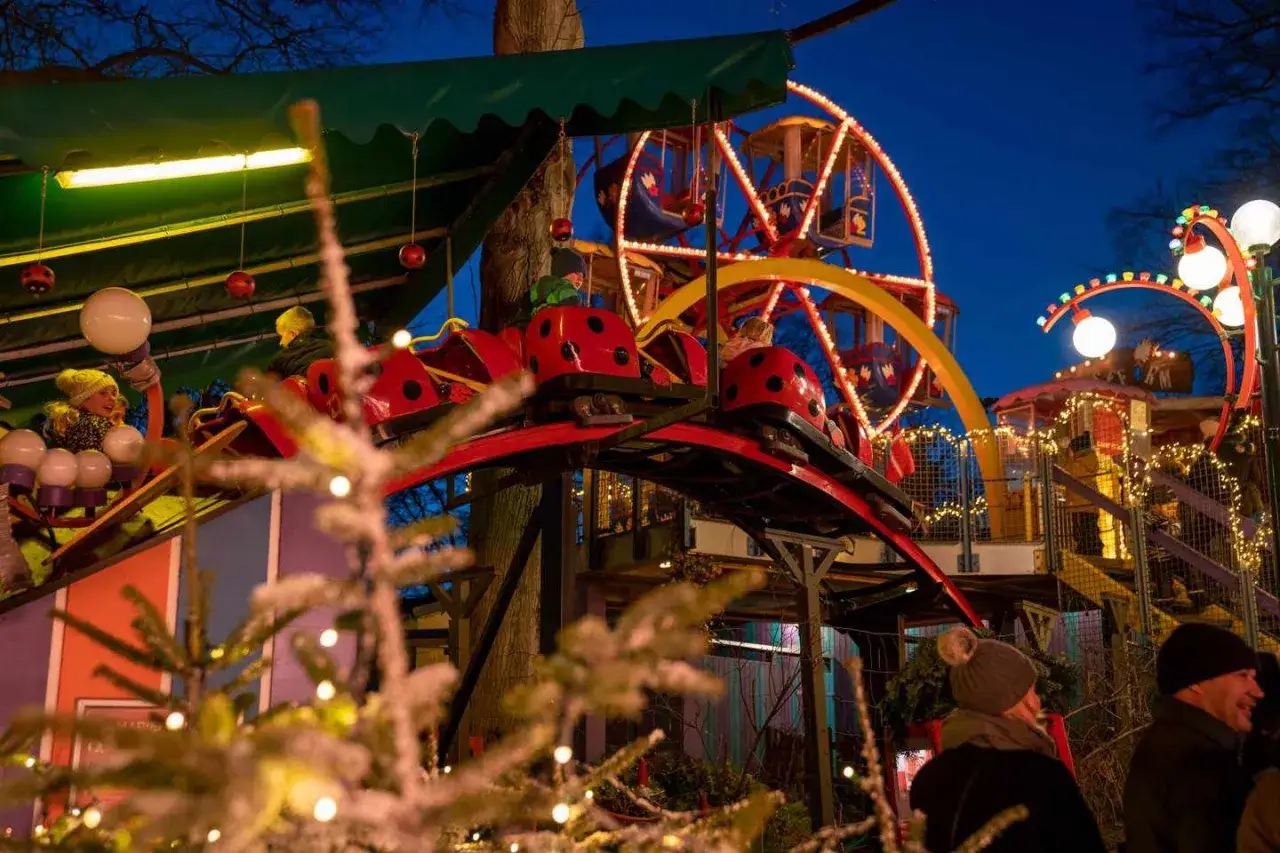 Barn njuter av en &aring;ktur p&aring; en nyckelpiga-berg-och-dalbana p&aring; en julmarknad. Ett par i f&ouml;rgrunden ler.