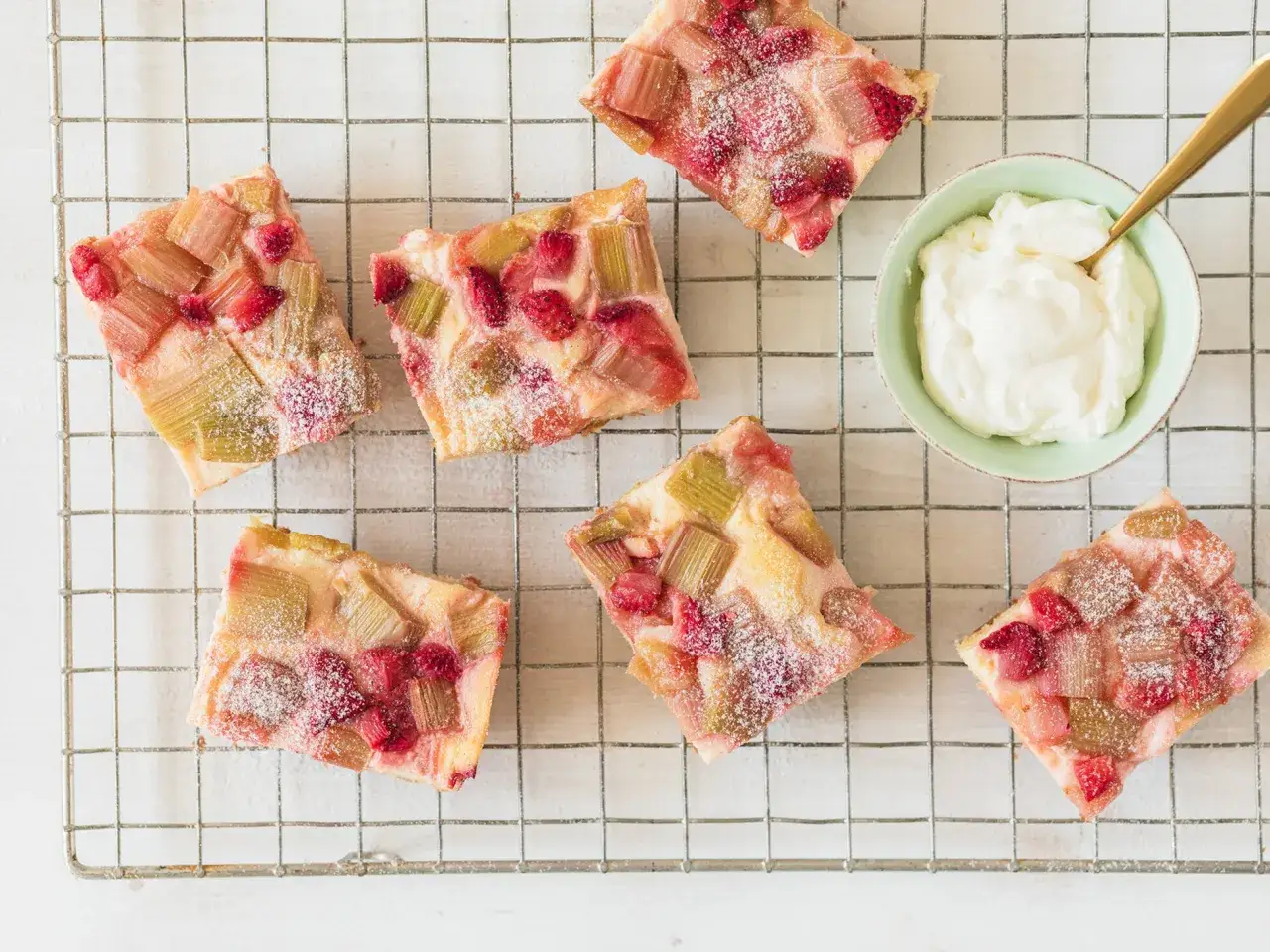 Saftiger Erdbeer Rhabarber Kuchen, best&auml;ubt mit Puderzucker, wartet neben einer Sch&uuml;ssel Schlagsahne auf seine Liebhaber.