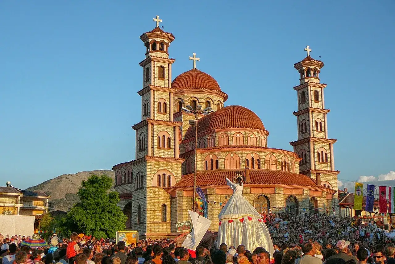 Albania ciekawostki: barwna procesja z postacią w sukni przed cerkwią. Tłumy ludzi na placu.