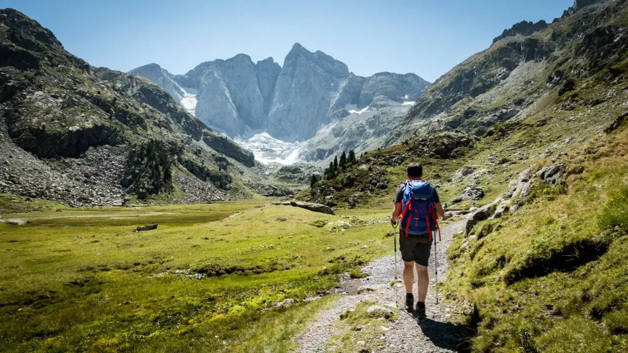 Un randonneur s'engage sur un sentier rocailleux, sac à dos bleu et rouge, prêt pour un trek pyrénéen. Le paysage montagneux s'étend, majestueux.