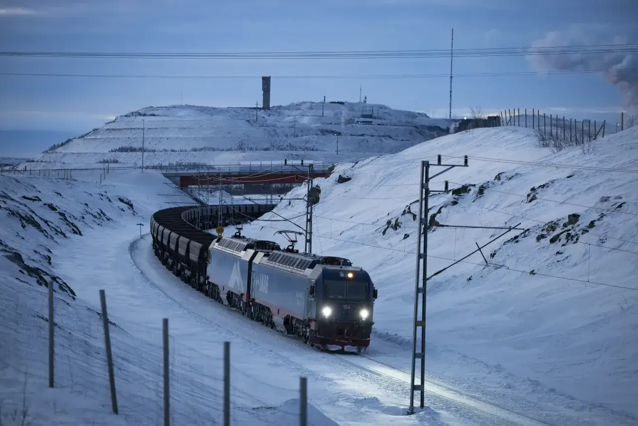 Ett långt godståg, ett tåg Luleå Narvik, slingrar sig fram genom ett snöigt landskap under en blågrå himmel.