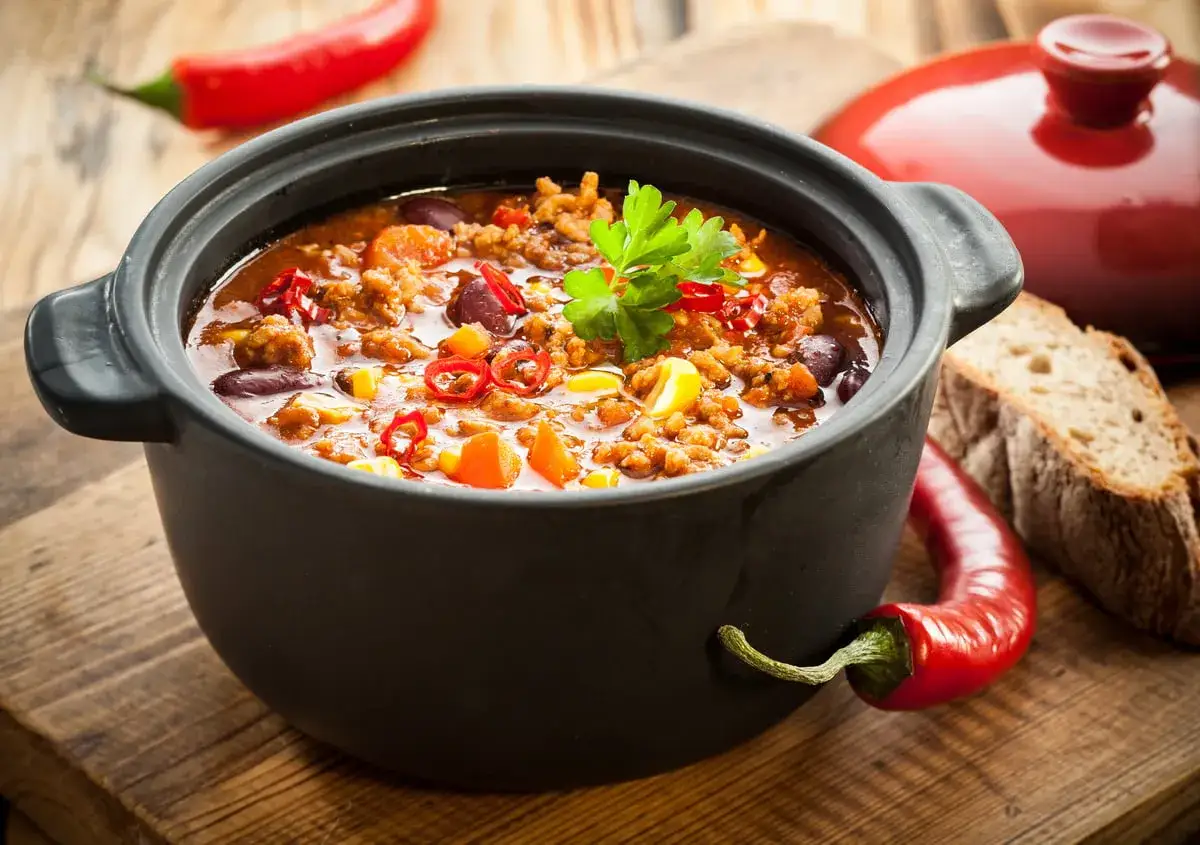 Un chili savoureux mijote dans une cocotte en fonte, parfaite pour aller au four. Accompagné de pain et de piments rouges.