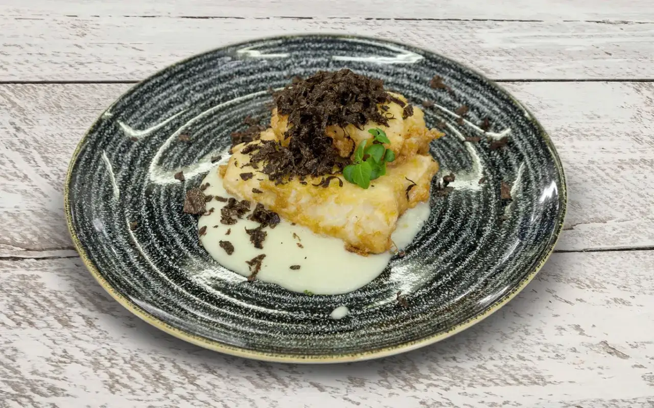Merluza a la romana crujiente, servida sobre una cremosa salsa blanca y coronada con virutas de trufa negra.