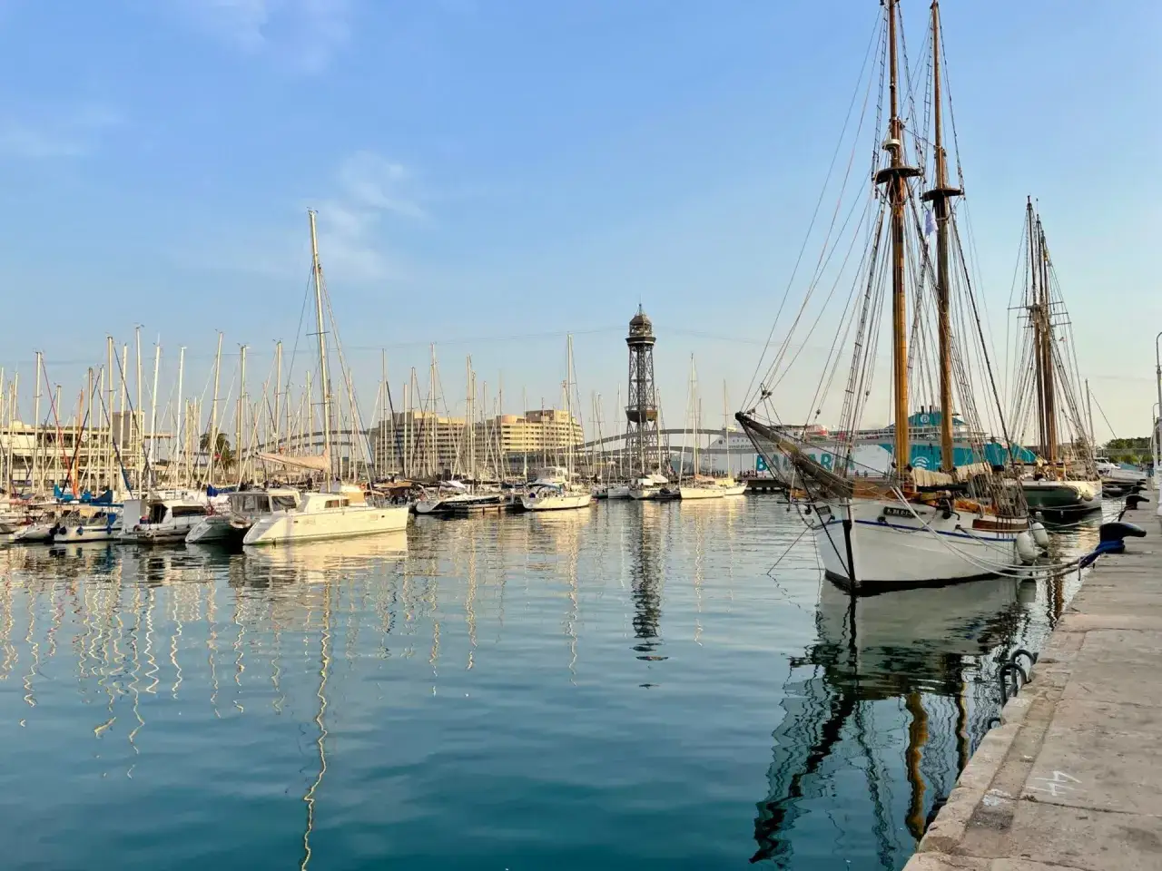 Port w Barcelonie, pełen żaglówek i statków, z wieżą widokową w tle. Czy w Barcelonie jest bezpiecznie? Tak, port wygląda na spokojny i bezpieczny.