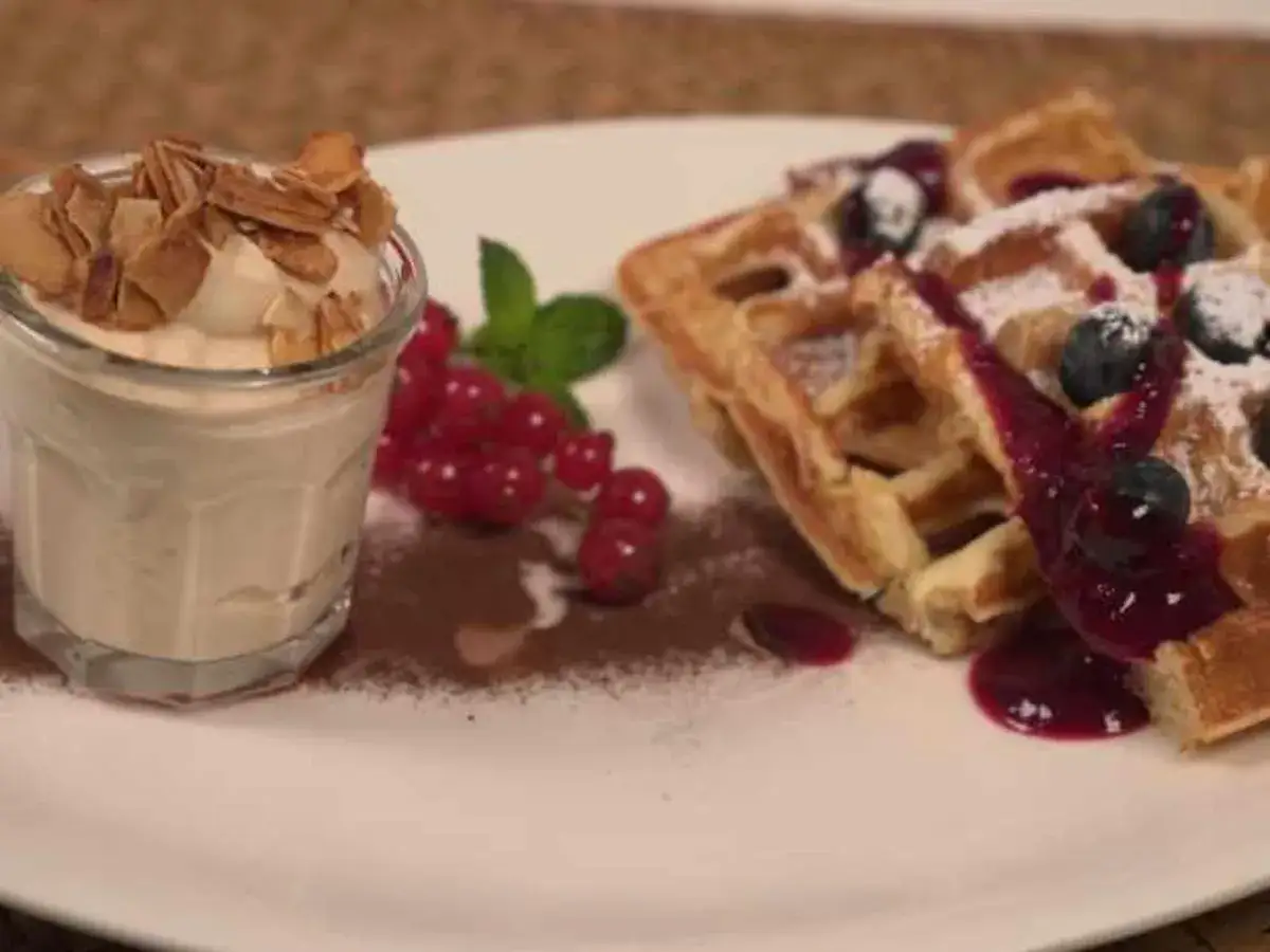 Belgische Waffeln mit Blaubeersauce, Puderzucker und einem Becher Creme mit Mandelsplittern.