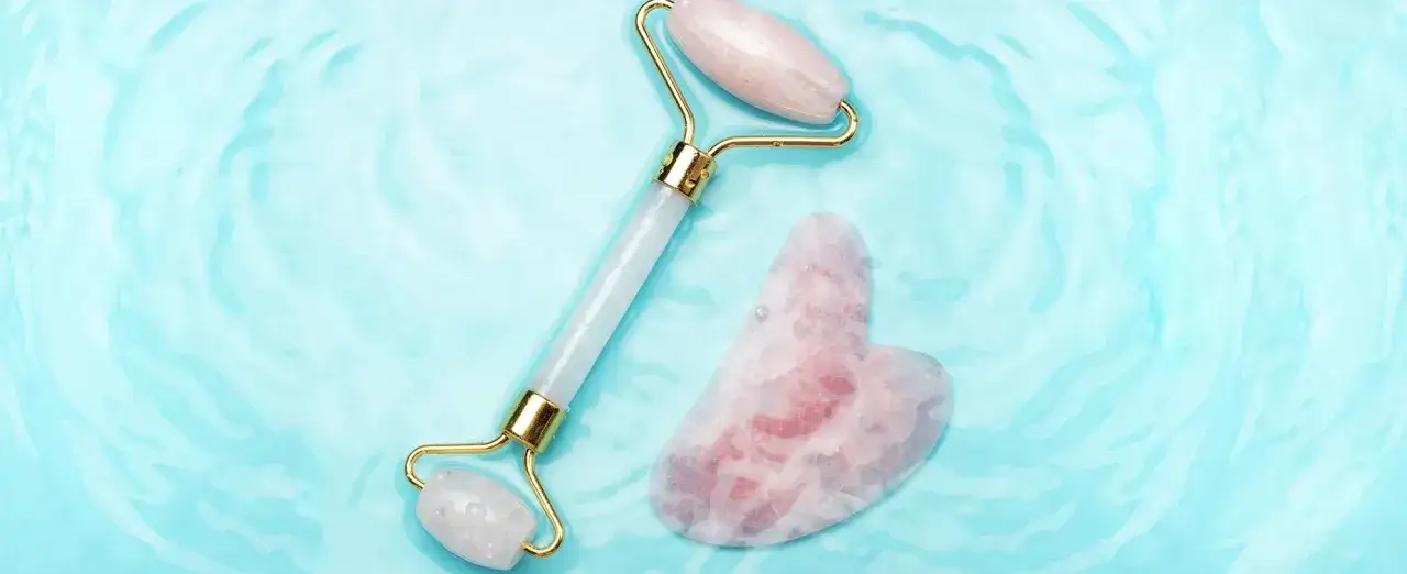 Un rullo di giada e una pietra gua sha a forma di cuore rosa su uno sfondo azzurro e ondulato, evocando una sensazione di freschezza e benessere.