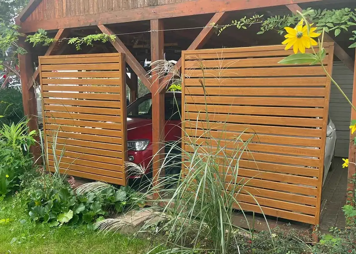 Holzlamellen verkleiden den Carport und schaffen eine moderne Optik. Gr&uuml;ne Pflanzen umrahmen die Einfahrt.