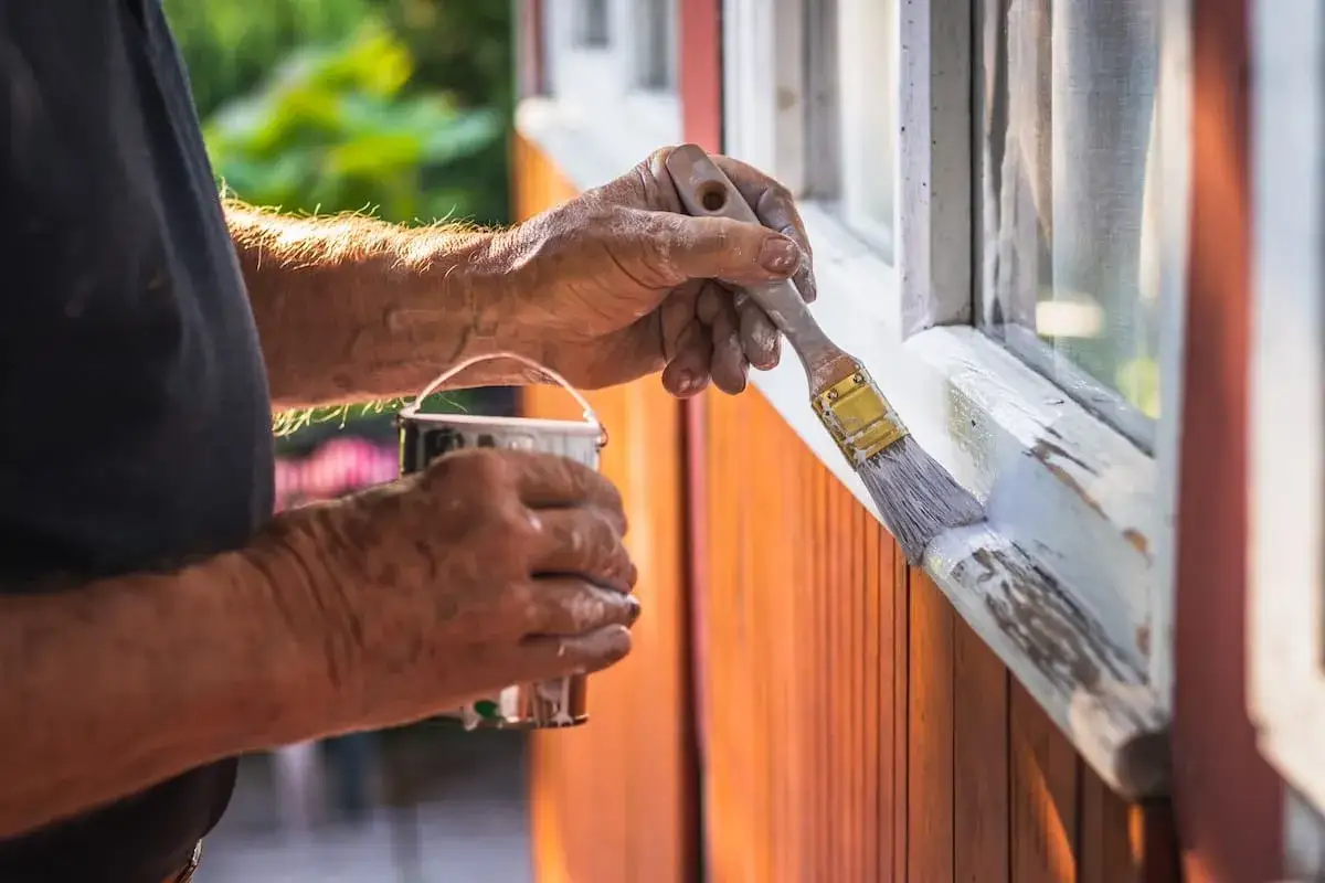 Mains d'un homme peignant du bois brut extérieur, protégeant la fenêtre avec une nouvelle couche de peinture blanche.