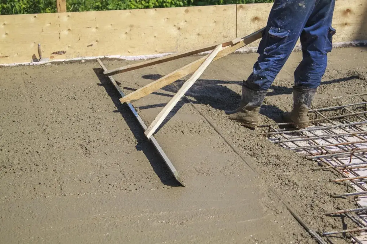 Pracownik wyr&oacute;wnuje świeżą wylewkę betonową za pomocą łaty. Dowiedz się, jak zrobić wylewkę betonową.