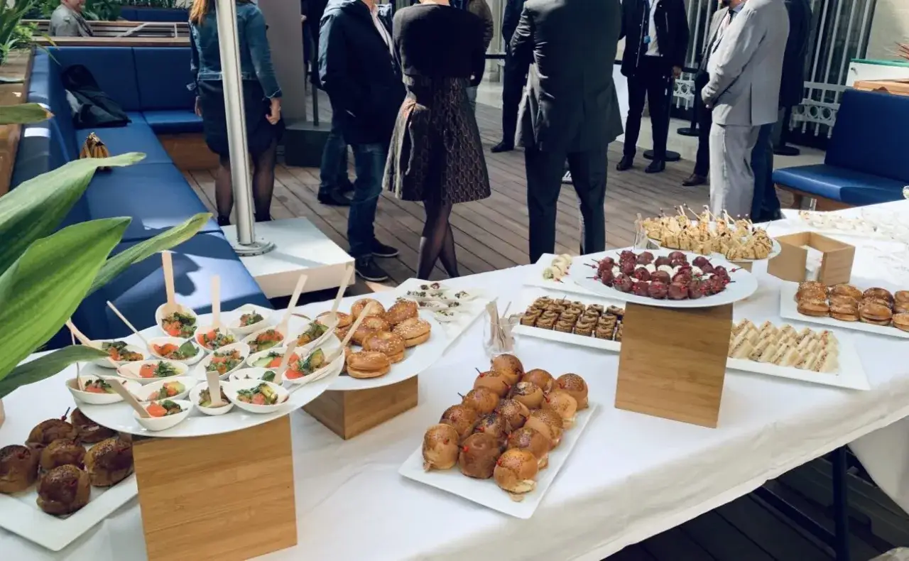 Un buffet froid avec des mini-burgers, des brochettes et des verrines de salade. Des invités discutent en arrière-plan.