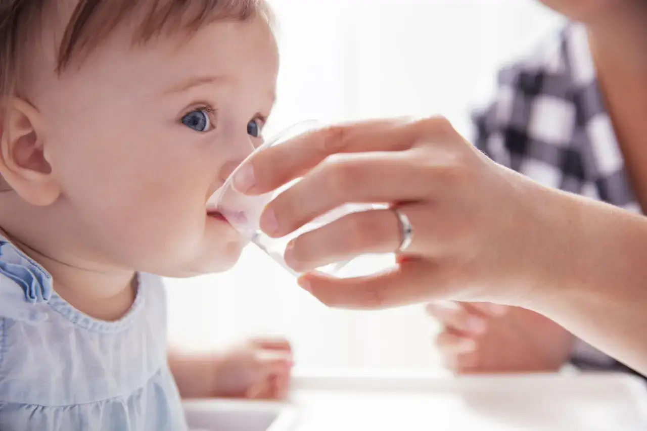 Ein Baby trinkt aus einem Becher, gehalten von einer Hand. Es scheint, als ob das baby trinkt zu viel, aber es ist nur ein kleiner Schluck Wasser.
