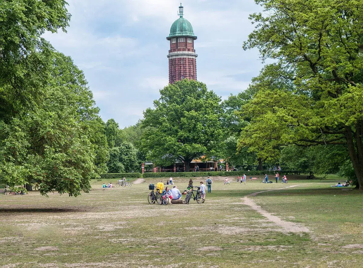 Ein sonniger Tag im Volkspark Jungfernheide. Menschen entspannen sich auf der Wiese, einige radeln, w&auml;hrend der markante Wasserturm im Hintergrund aufragt.