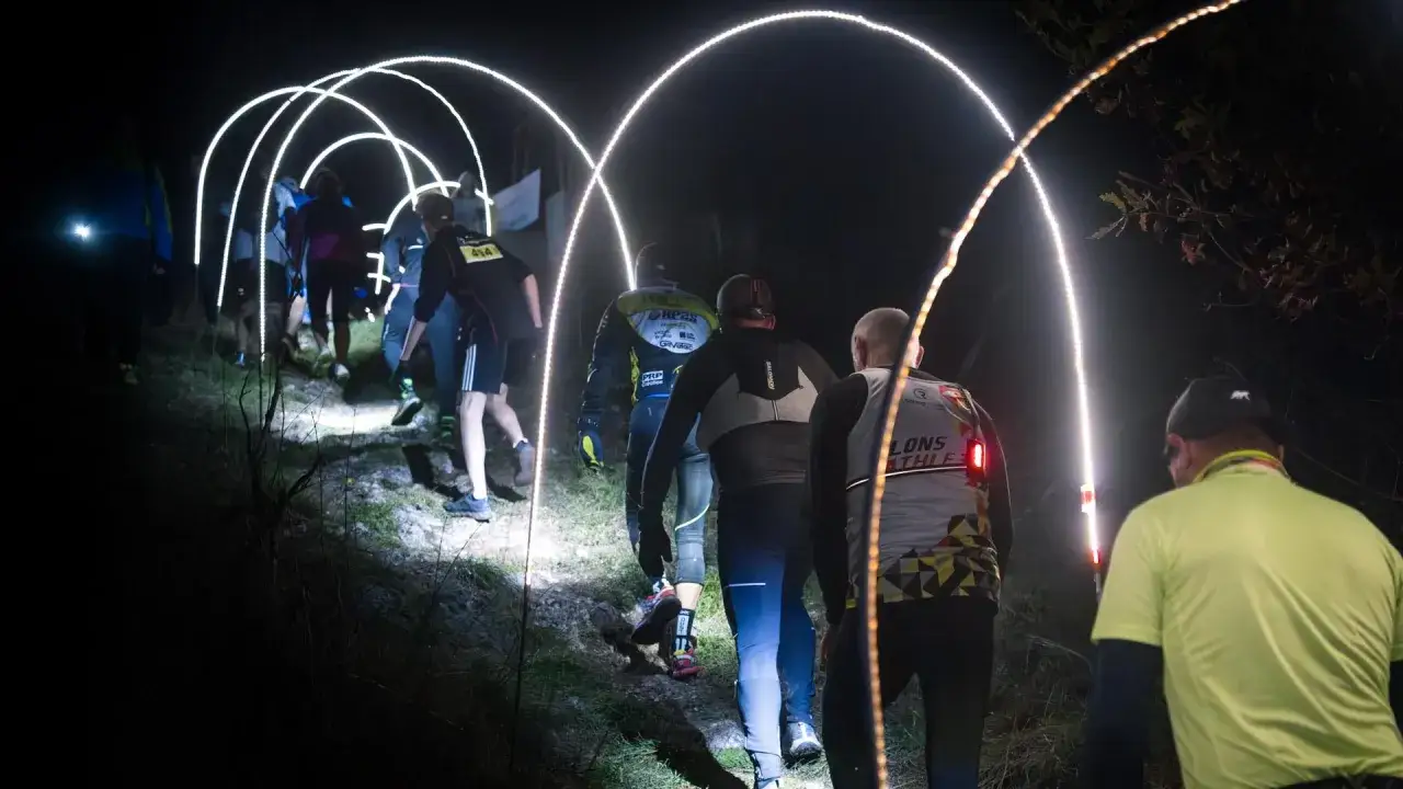 Coureurs dans un **dark trail** à Orgelet, éclairés par des arches lumineuses sur un sentier nocturne.