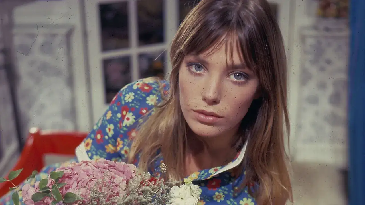 Jane Birkin z charakterystyczną fryzurą birkin bangs, w kwiecistej sukience, z bukietem różowych kwiatów.