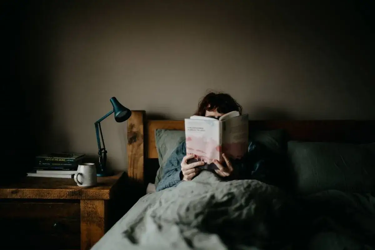 Persona leyendo libros para dormir en la cama, con una l&aacute;mpara de noche y una taza en la mesita.