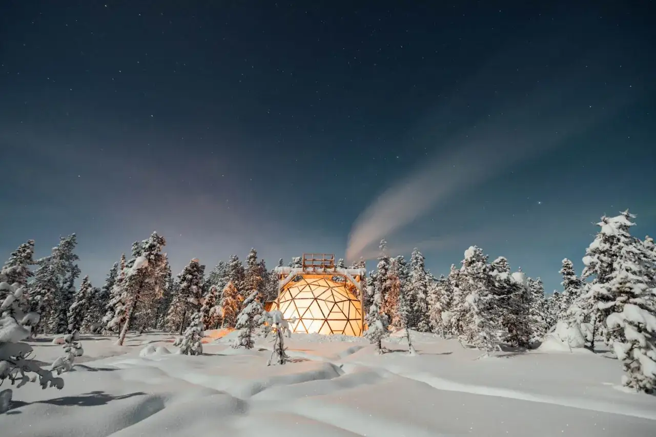 Un d&ocirc;me lumineux au c&oelig;ur d'un paysage laponie hiver, entour&eacute; de sapins enneig&eacute;s sous un ciel &eacute;toil&eacute;. La fum&eacute;e s'&eacute;chappe d'une chemin&eacute;e, cr&eacute;ant une atmosph&egrave;re f&eacute;erique.