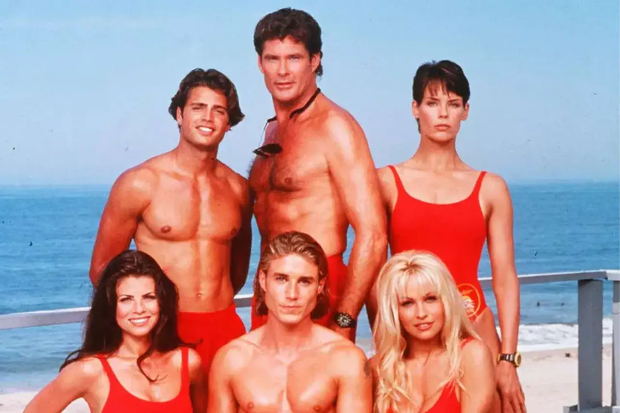 Słoneczny patrol obsada: David Hasselhoff, Pamela Anderson i inni ratownicy w czerwonych strojach kąpielowych pozują na plaży.