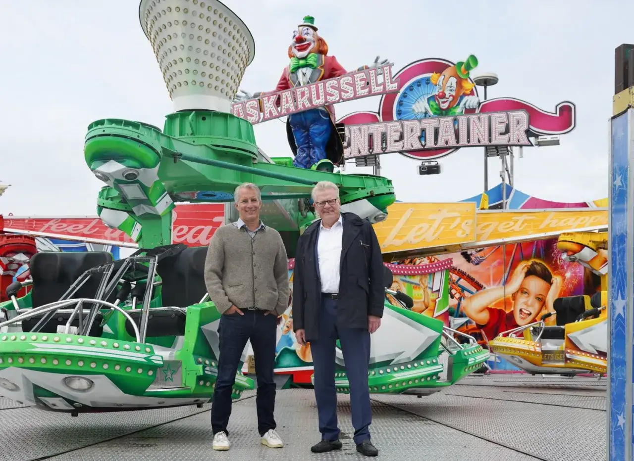 Zwei M&auml;nner stehen vor einer bunten Kirmesattraktion mit Clowns und der Aufschrift 