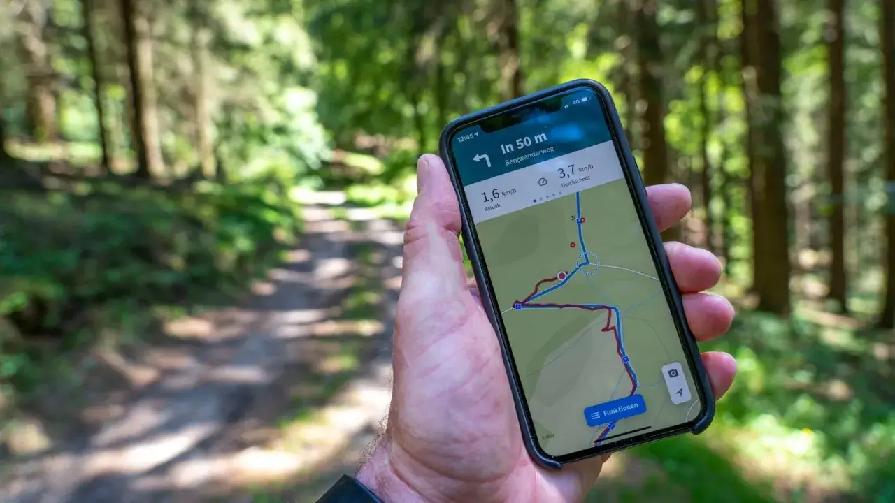 Hand h&auml;lt Smartphone mit komoot Wanderwege-App im Wald. Die Karte zeigt eine Route auf einem Bergwanderweg.