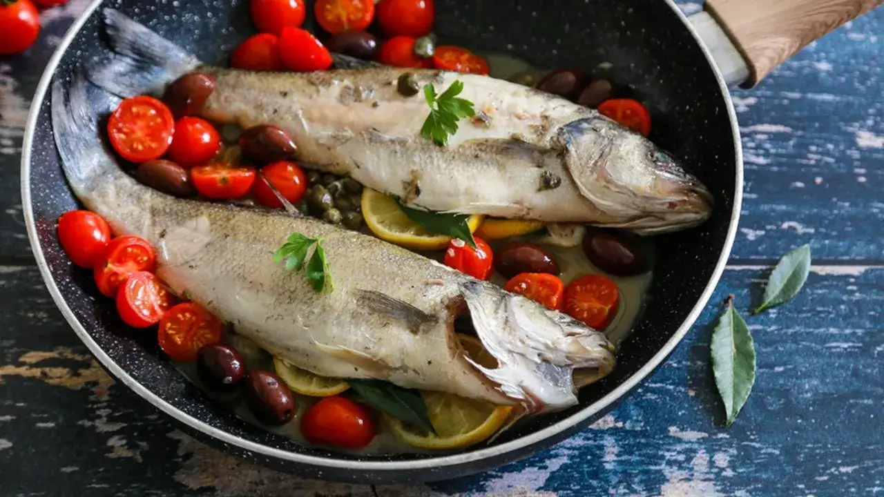 Due branzini cotti in padella con pomodorini, olive, limone e prezzemolo. Un piatto saporito e colorato.