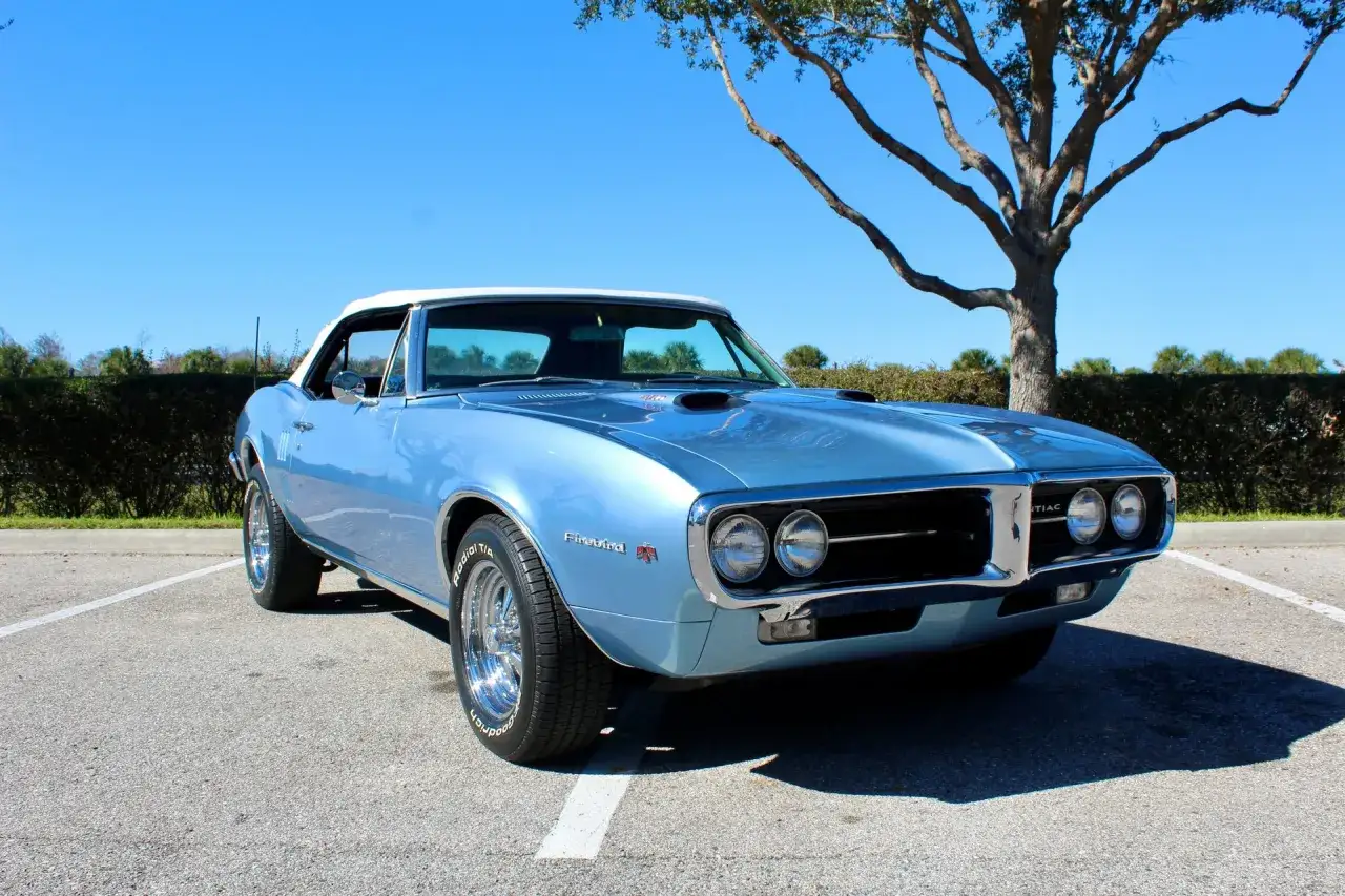 Bl&aring; cabriolet Pontiac Firebird 69 parkerad under ett tr&auml;d med klarbl&aring; himmel.