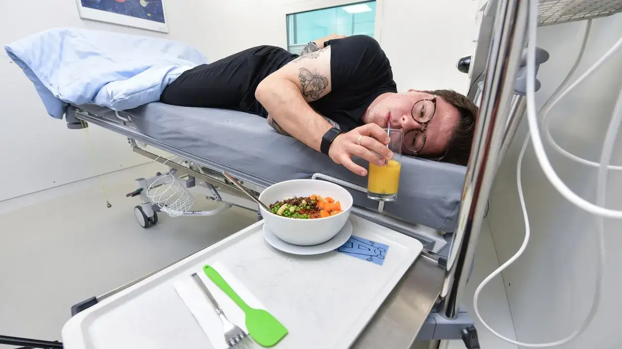 Mann liegt auf einem Krankenbett, trinkt Saft und isst eine Mahlzeit. Ein **bedside test** könnte hier stattfinden.