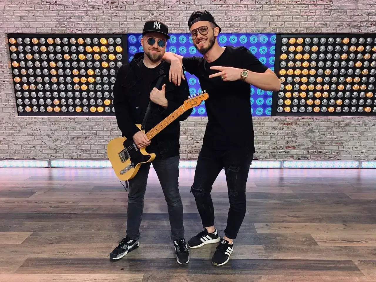 Dwóch łobuzów członków zespołu, jeden z gitarą, drugi pozuje z gestem pokoju, na tle ściany z cegły i świateł.