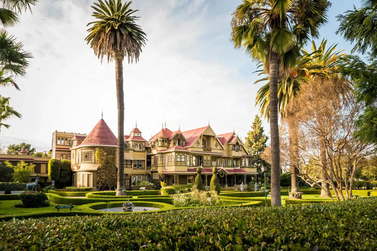La casa Winchester, una mansión victoriana de color amarillo con tejados rojos, rodeada de palmeras y jardines cuidados.