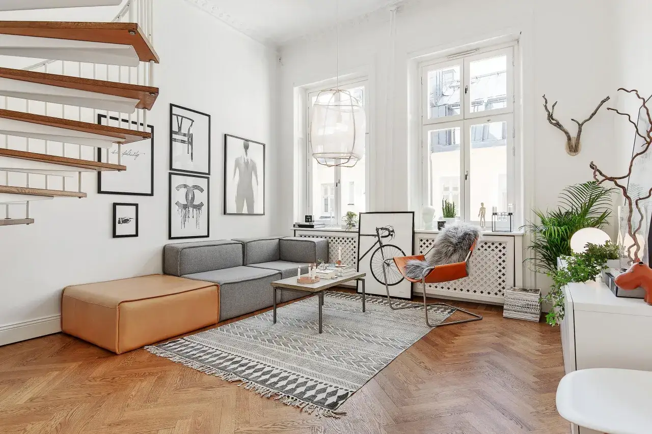 En modern lägenhet med en grå soffa, orange fåtölj och en stor matta. En trappa leder uppåt. Konst på väggarna och växter ger liv åt rummet, en hyllning till svenska designklassiker.