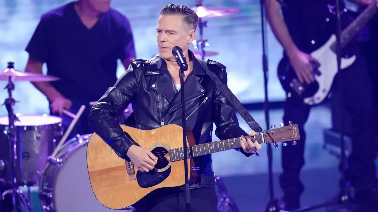 Bryan Adams na koncercie w sk&oacute;rzanej kurtce gra na gitarze akustycznej, w tle zesp&oacute;ł.