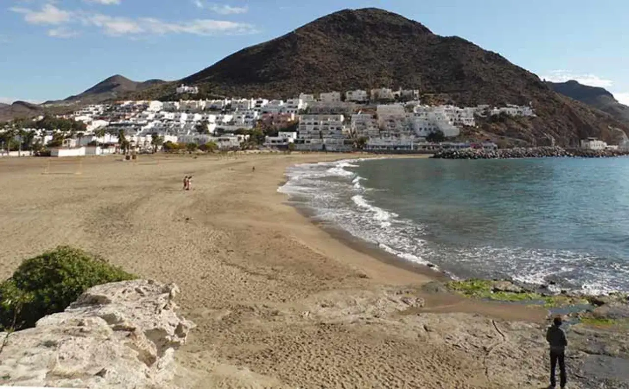 Amplia playa de arena dorada en la playa de San José, con el mar azul y un pueblo blanco al fondo, bajo una montaña.