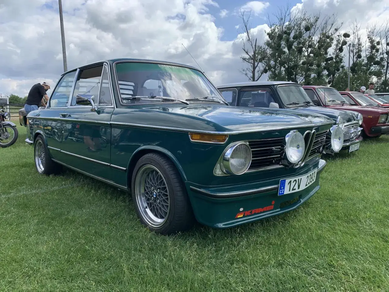 Klasyczne BMW 2002 w kolorze ciemnozielonym, zaparkowane na trawie obok innych samochodów.