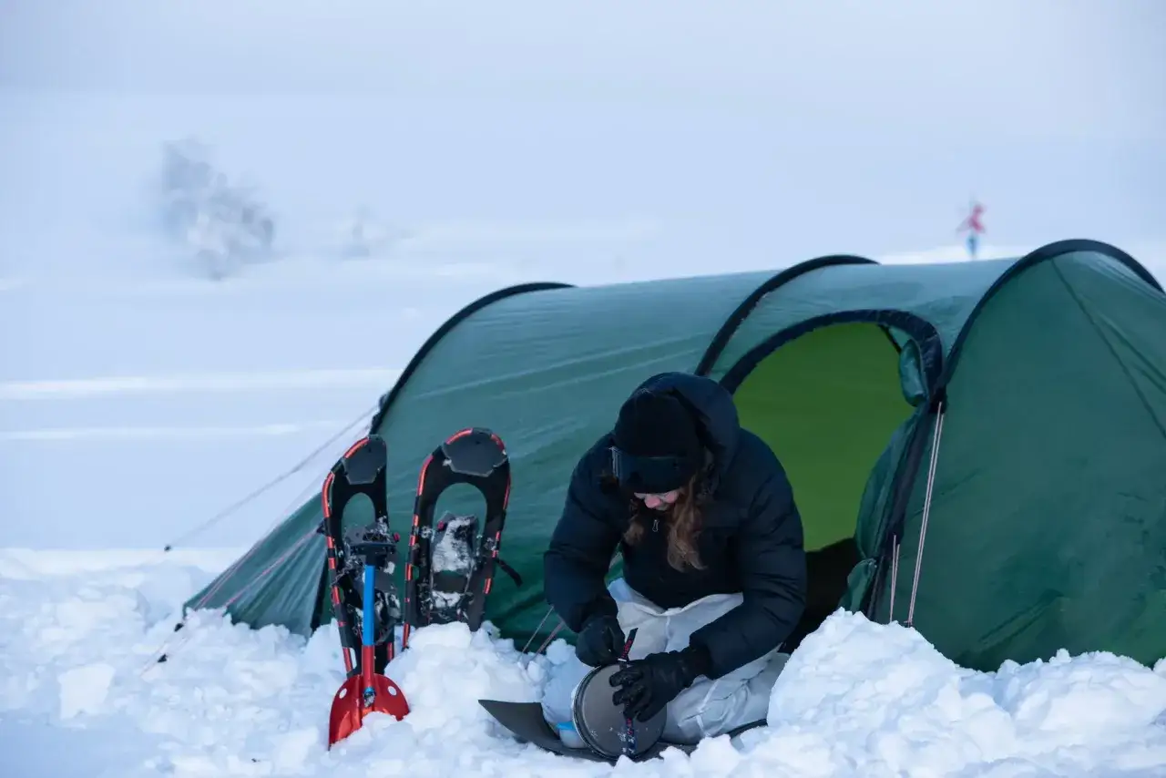 Person f&ouml;rbereder mat vid ett t&auml;lt i sn&ouml;n, med sn&ouml;skor och spade redo. Snart blir det v&auml;rme i bilen p&aring; vintern.