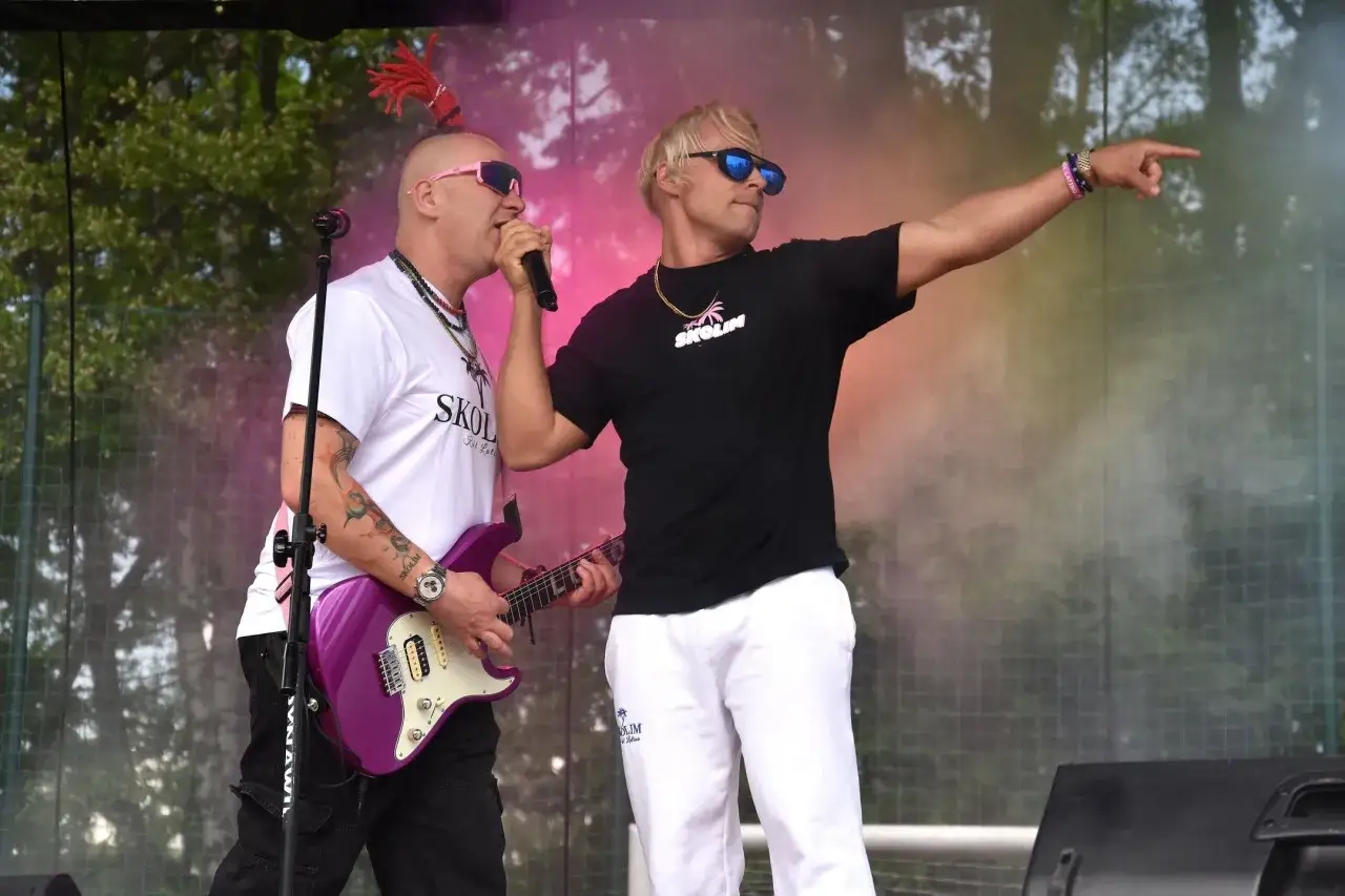 Dwóch mężczyzn na scenie, jeden z gitarą, drugi wskazuje palcem. Na koszulkach logo