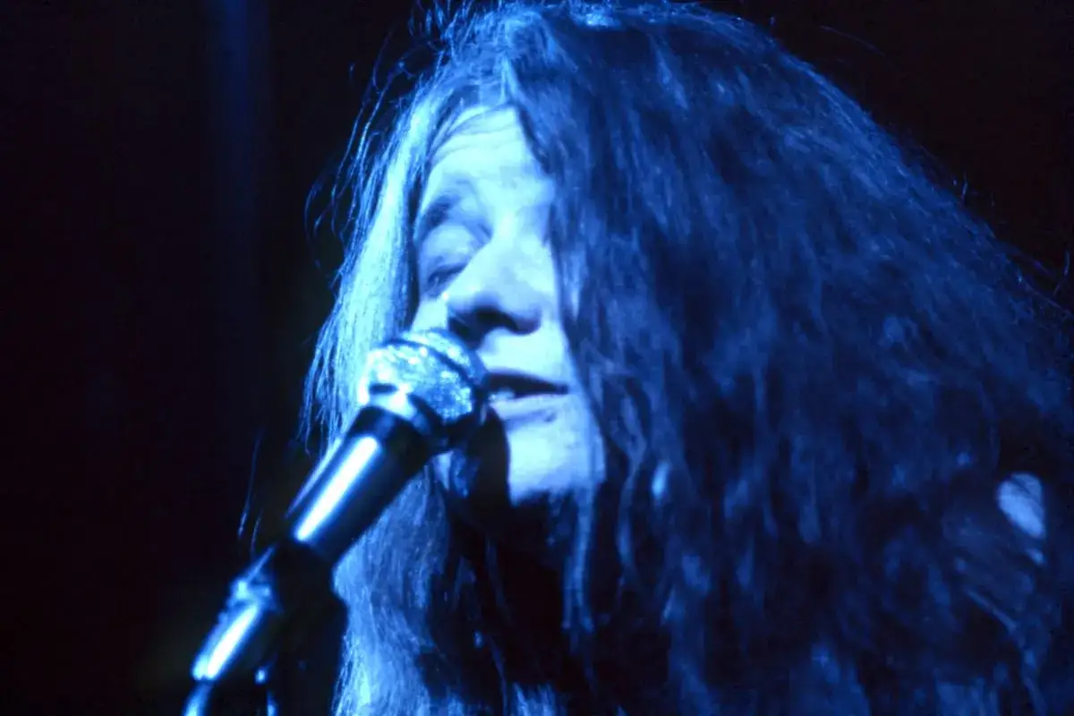 Janis Joplin, con su melena suelta y voz potente, canta apasionadamente al micrófono bajo una luz azul.