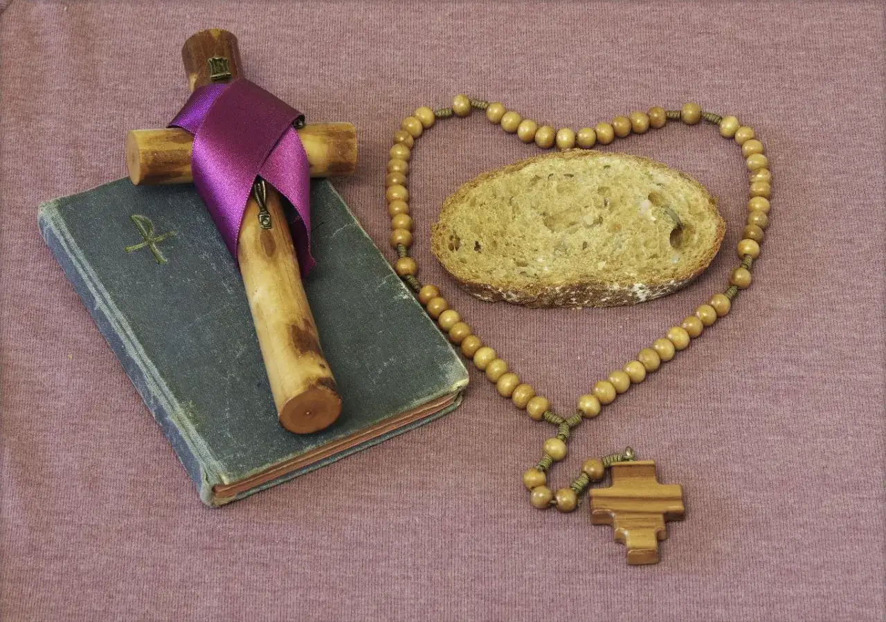 Krzyż, książka i bochenek chleba tworzą symboliczne tło do pytania: czy w Wielki Piątek można jeść mięso? Różaniec w kształcie serca otacza kromkę.