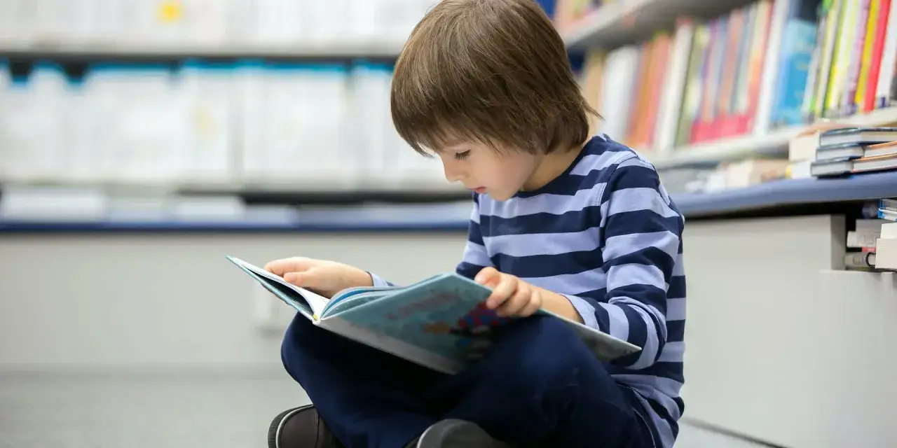 Chłopiec w pasiastej bluzce z zapałem ćwiczy czytanie ze zrozumieniem klasa 2, siedząc na podłodze w bibliotece.