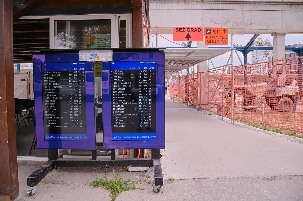 Anzeigetafeln am Ljubljana Bahnhof zeigen Zugabfahrten und -ank&uuml;nfte. Baustellenabsperrungen und Bauarbeiten sind sichtbar.