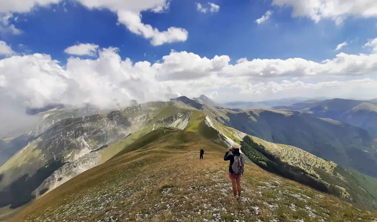 Persone in cima a un crinale erboso, con vista panoramica sulle montagne dei Monti Sibillini. Il cielo &egrave; azzurro con nuvole bianche soffici.