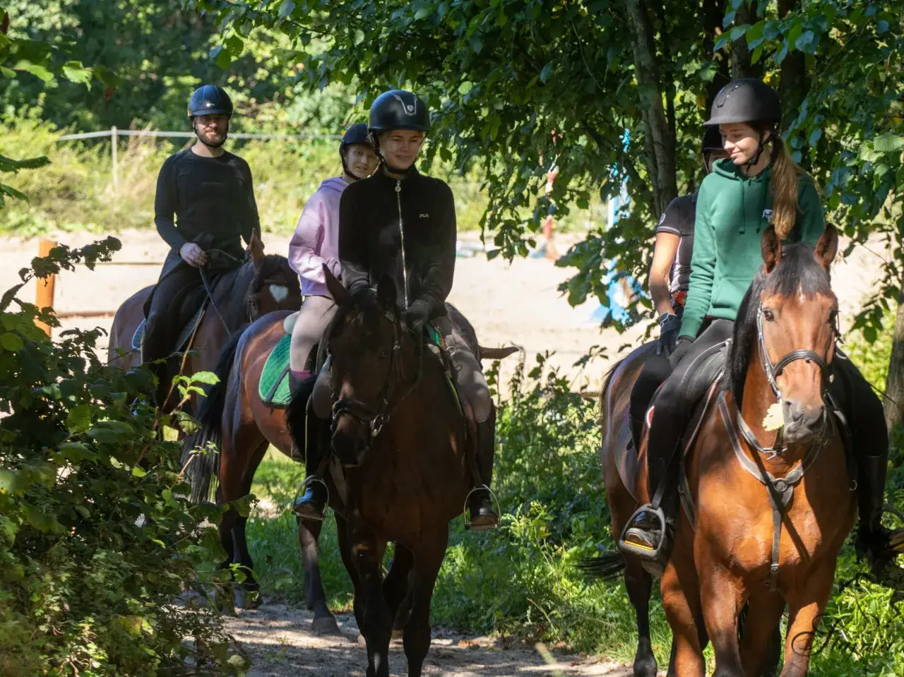 Grupa jeźdźców na koniach podczas przejażdżki w lesie. Wszyscy mają kaski. Klub jazdy konnej Fabrex zaprasza na wspólne wyprawy.