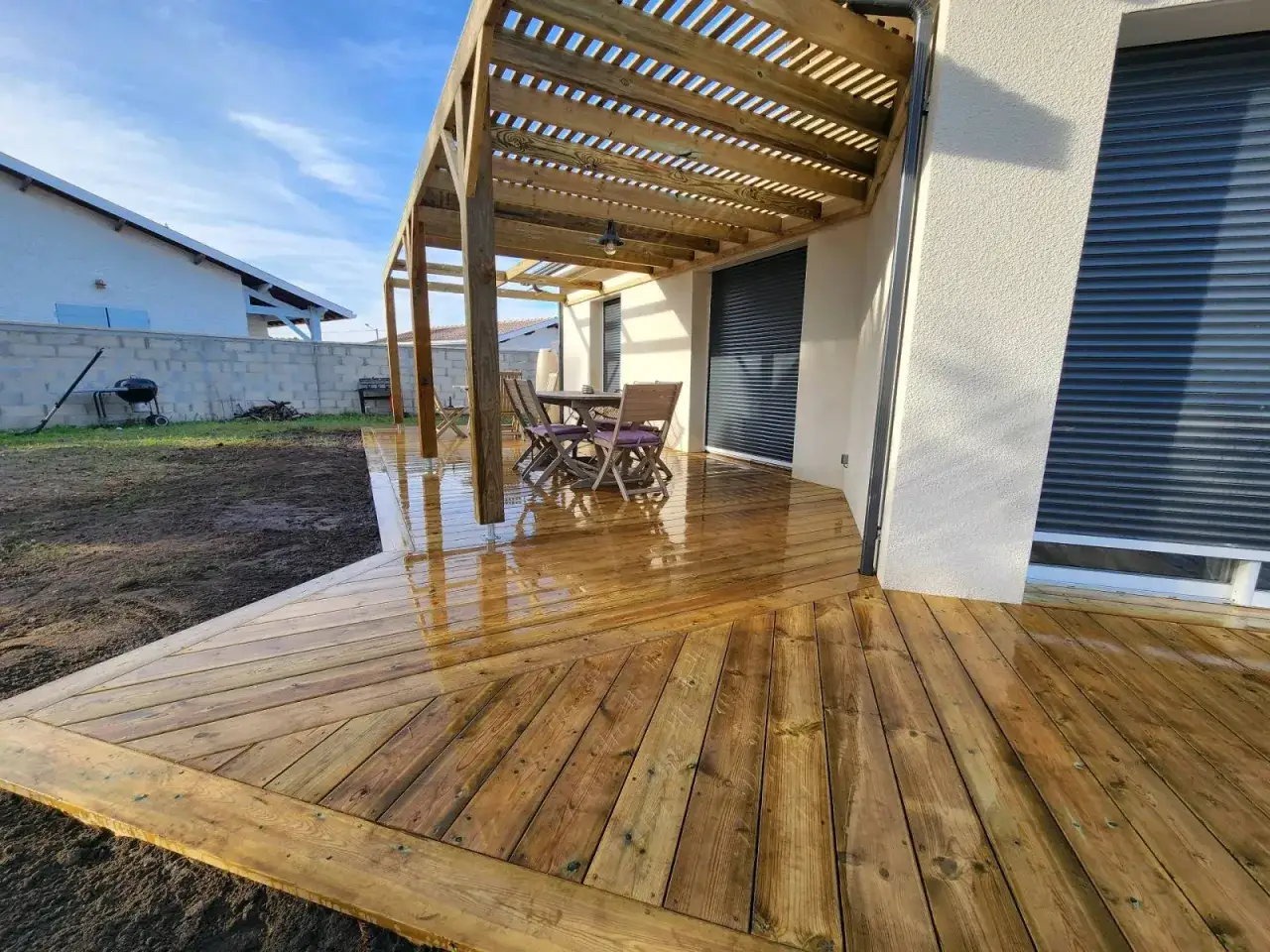 Terrasse en bois classe 3 fraîchement traitée, reflétant le ciel bleu. Une table et des chaises attendent sous la pergola.