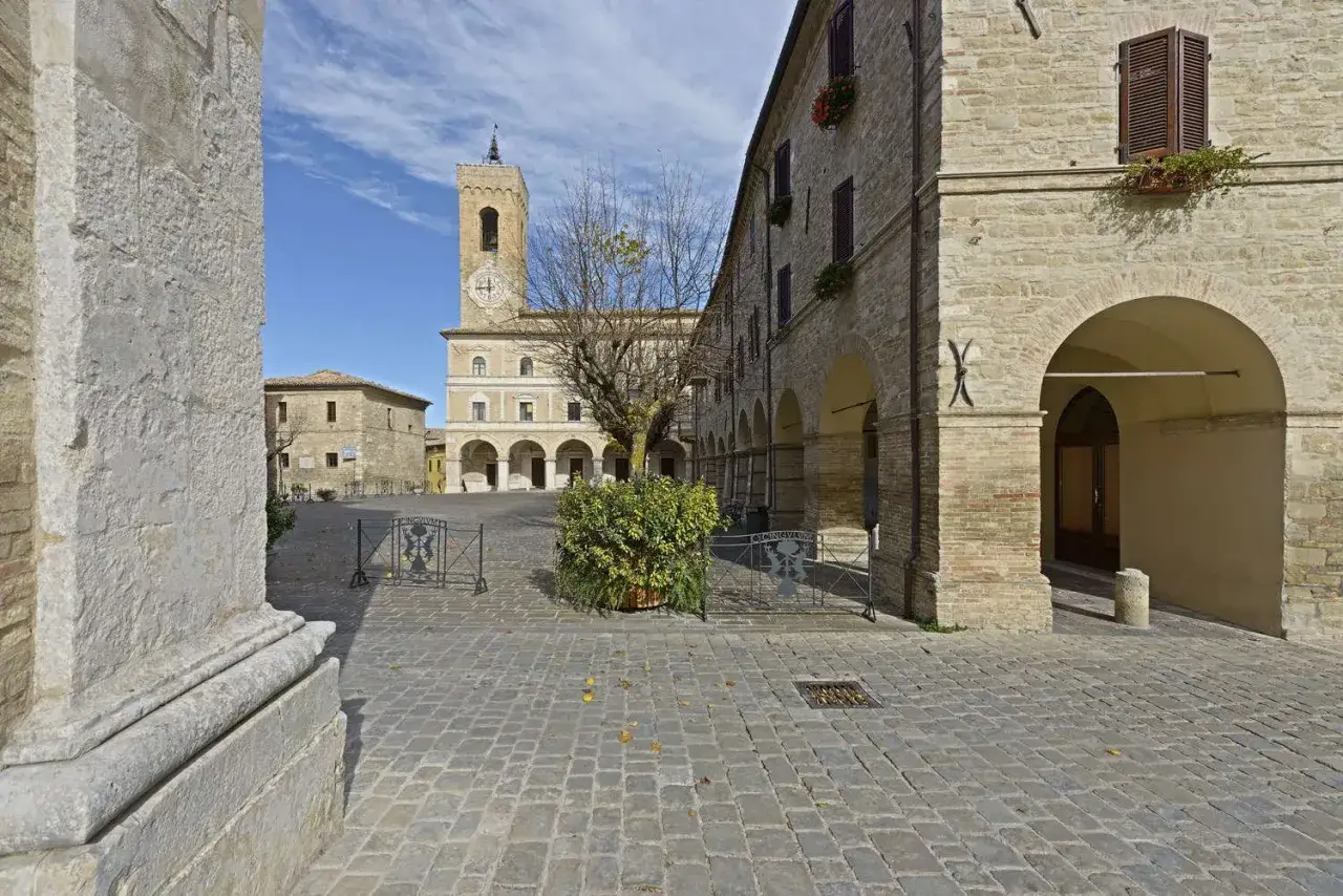 Place pavée de **Cingoli** avec une tour d'horloge imposante, des arcades et des bâtiments en pierre sous un ciel bleu.