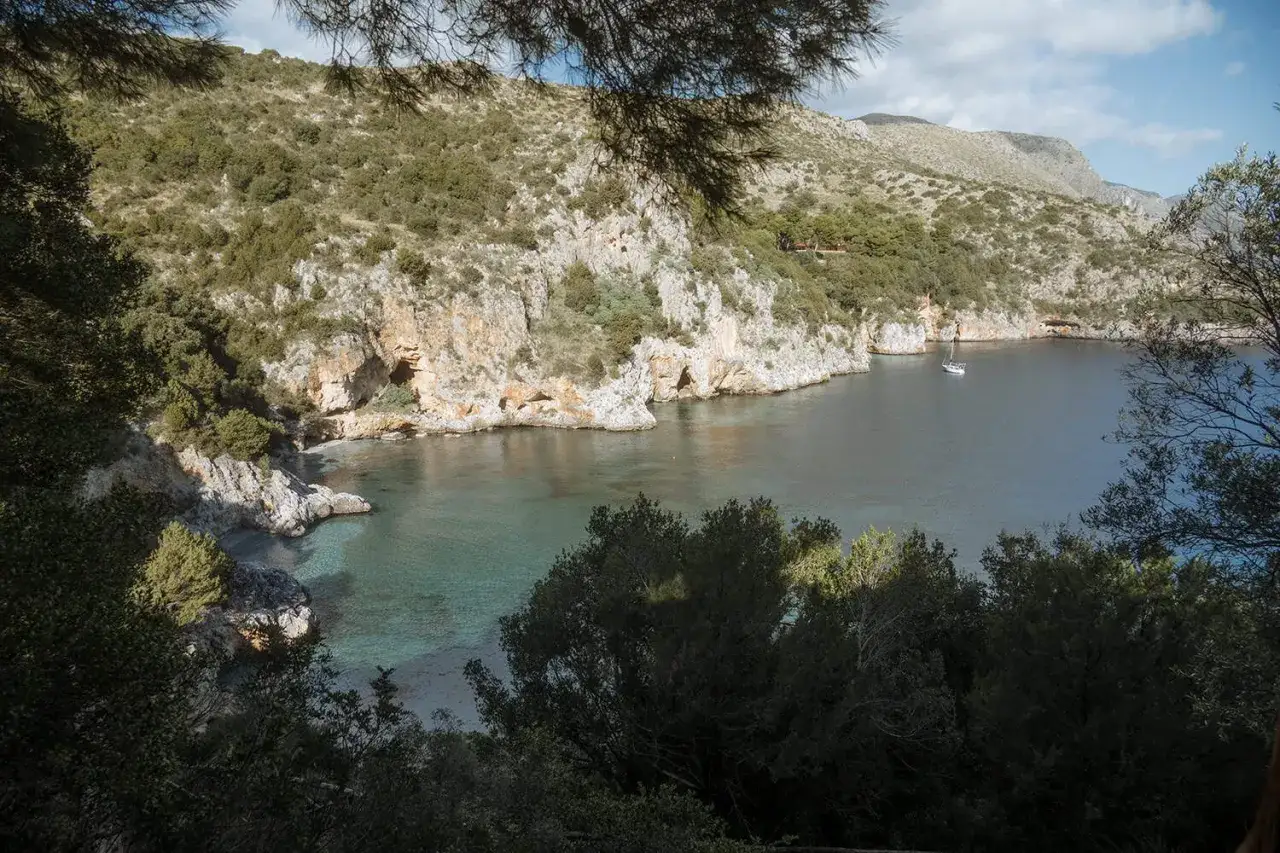 A secluded cove, Baia degli Infreschi, with clear turquoise waters, rocky cliffs, and lush green vegetation. A lone sailboat drifts peacefully in the bay.