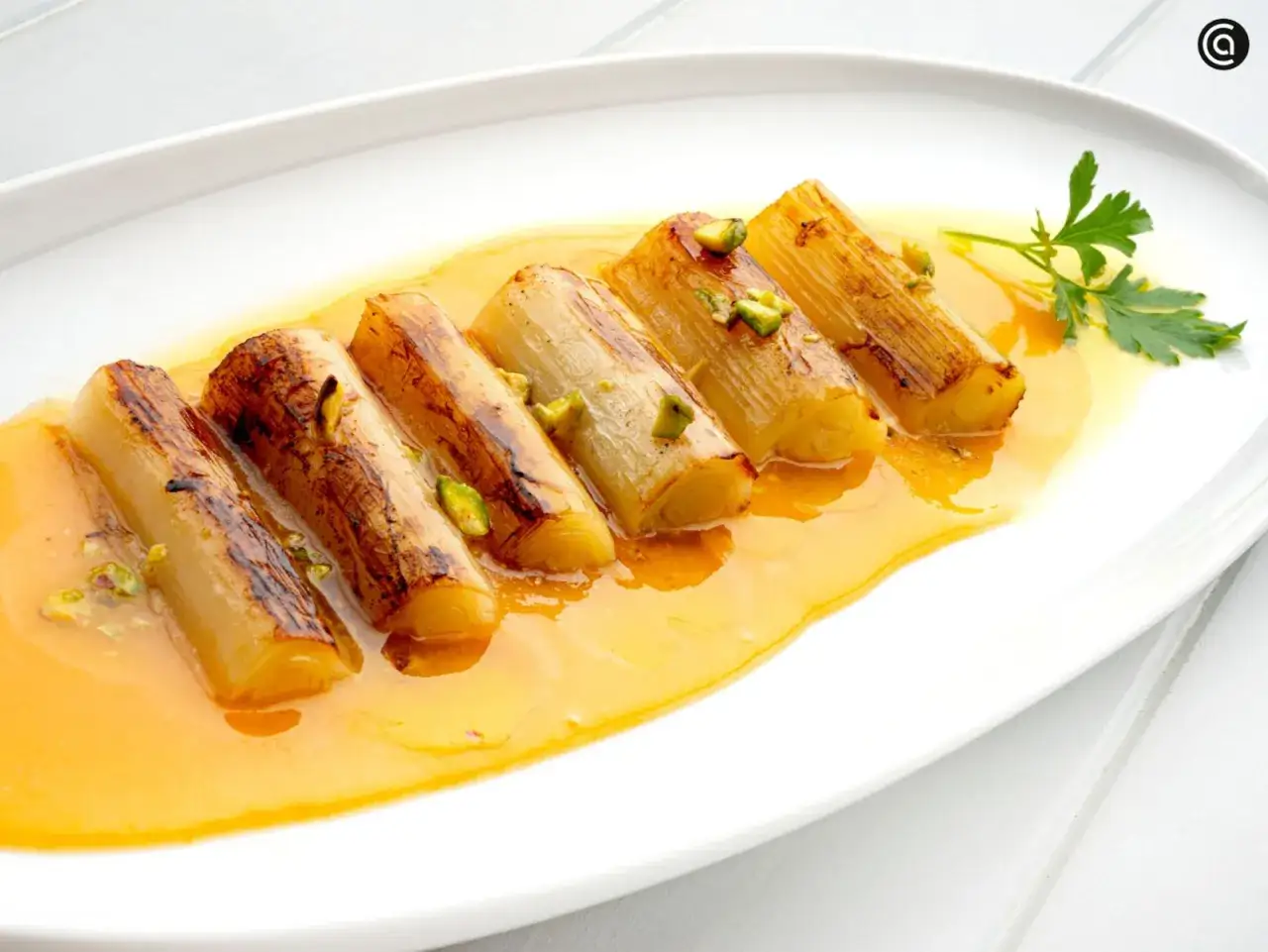 Puerros a la plancha caramelizados, servidos en una salsa dorada y espolvoreados con pistachos picados.