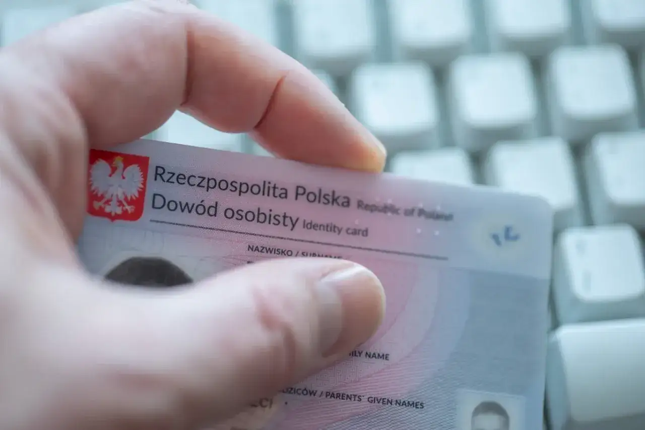 Dłoń trzyma polski dowód osobisty na tle klawiatury. Dokument zawiera godło i napis 