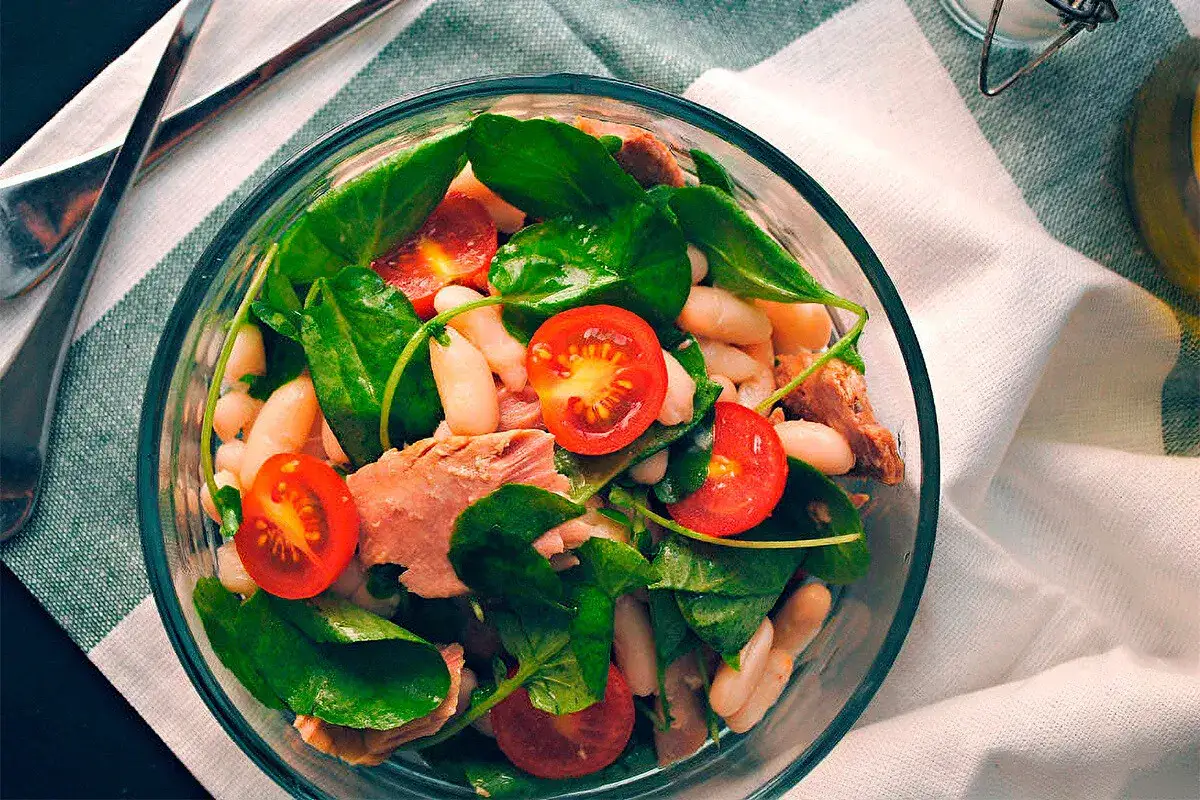 Ensalada de alubias con atún, espinacas y tomates cherry, servida en un bol de cristal.