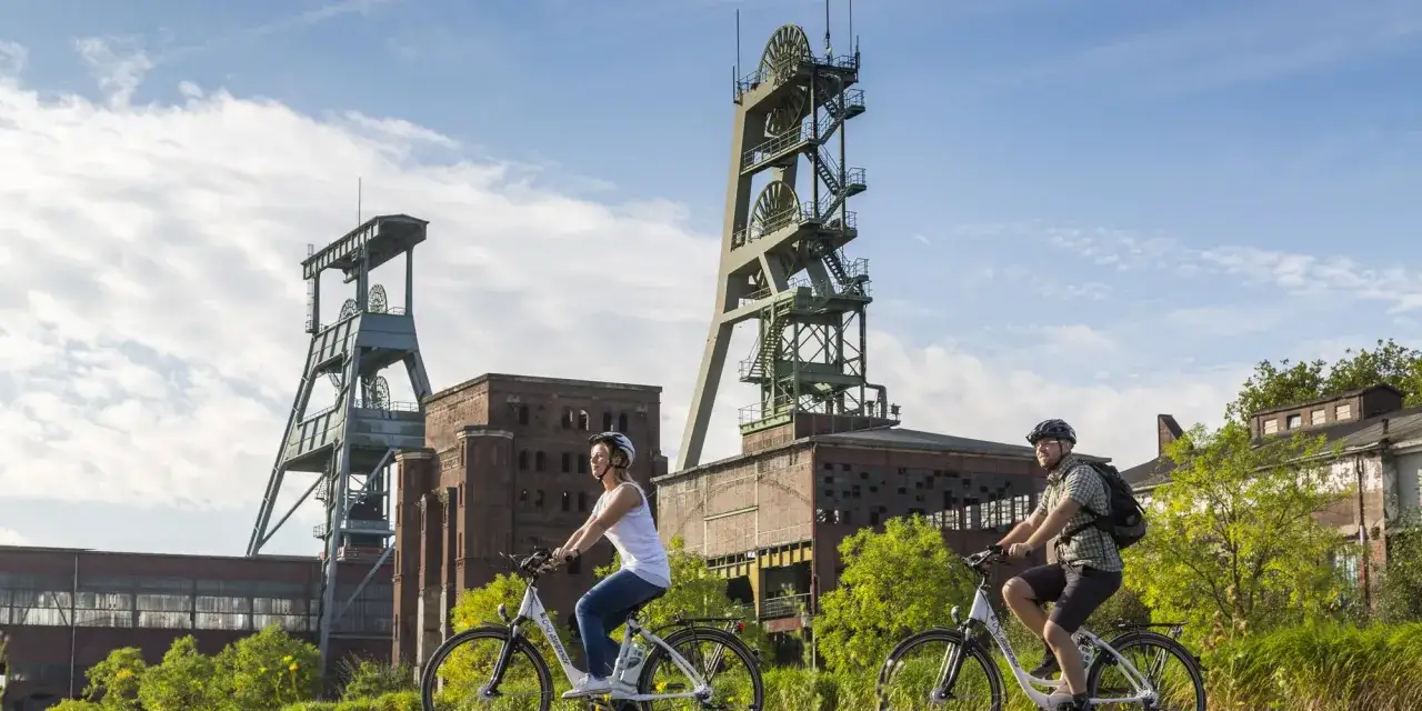 Zwei Radfahrer genießen die Landschaft am ehemaligen Bergwerk Ewald. Die imposanten Fördertürme stehen im Hintergrund.