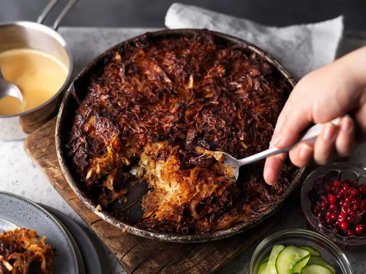 En gyllene, krispig farmors k&aring;lpudding i en form, med lingon och gurka vid sidan. En sked tar en portion.