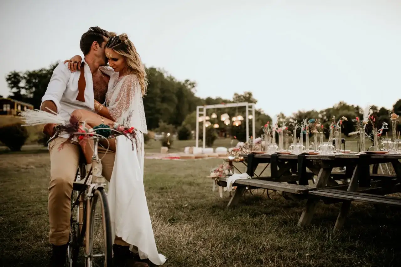 Un couple s'embrasse sur un v&eacute;lo d&eacute;cor&eacute;, devant une table dress&eacute;e pour un mariage champ&ecirc;tre.