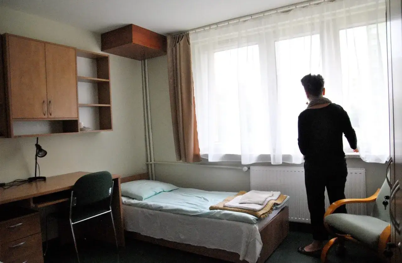 Pokój w akademiku w Lublinie: łóżko, biurko, szafa i osoba patrząca przez okno.